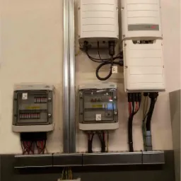 Szare skrzynki z bezpiecznikami i falowniki z okablowaniem zamontowane na ścianie wewnątrz budynku, widoczne przewody w peszlach i listwa montażowa.