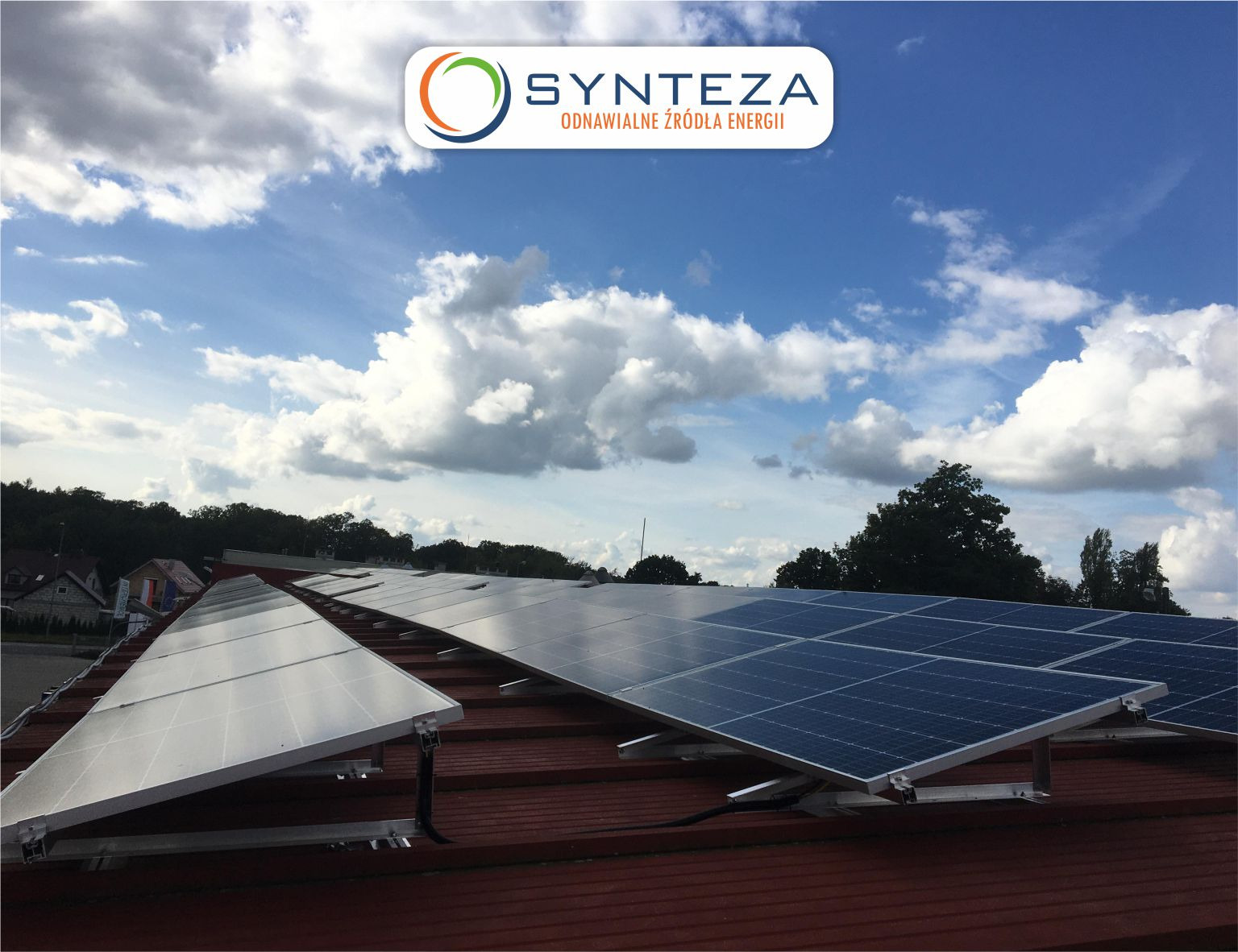Instalacja paneli na dachu z widocznym logo firmy na tle błękitnego nieba z chmurami.