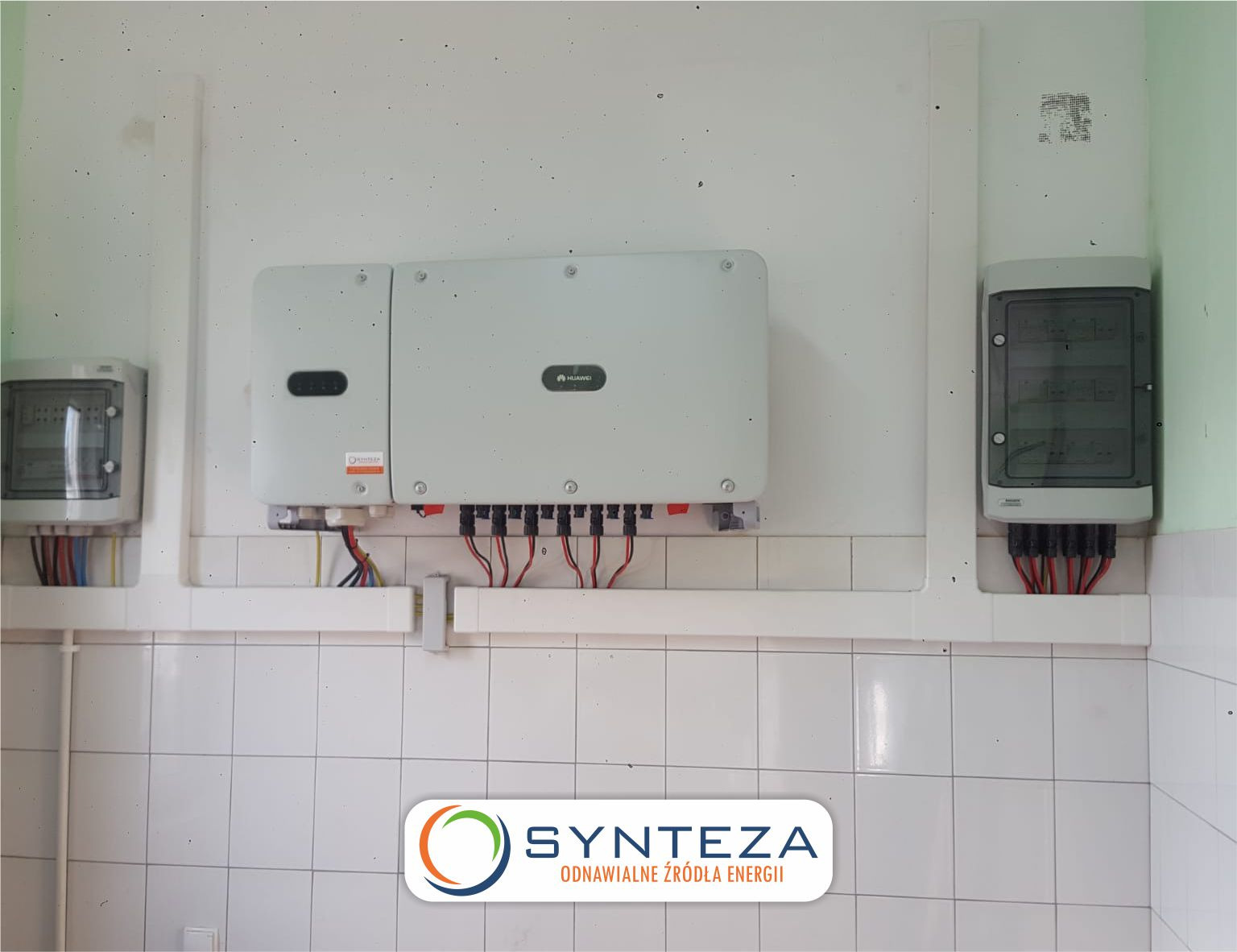 Zamontowany inwerter Huawei z okablowaniem i skrzynkami zabezpieczającymi na ścianie wyłożonej białymi, kwadratowymi płytkami. Widoczne logo firmy Synteza Odnawialne Źródła Energii.