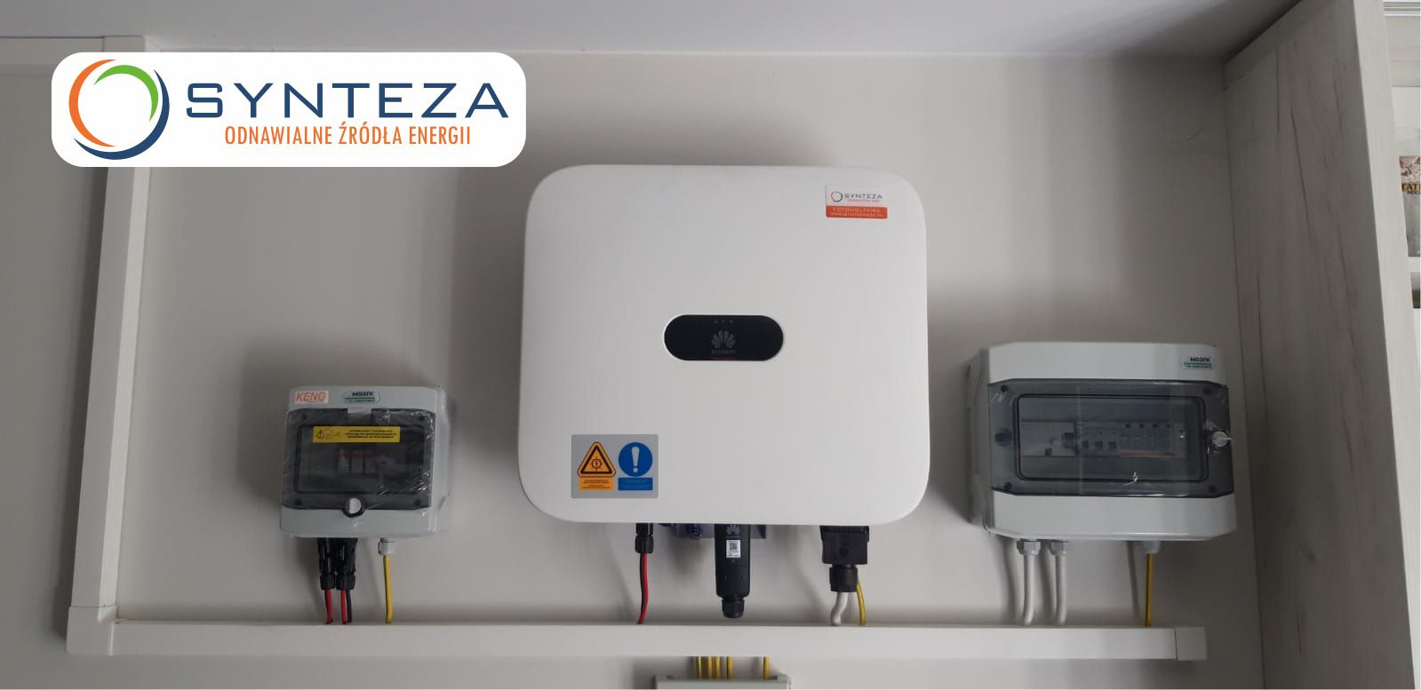 Biały falownik Huawei zamontowany na ścianie, obok dwie skrzynki elektryczne z przewodami. Widoczne logo firmy Synteza.