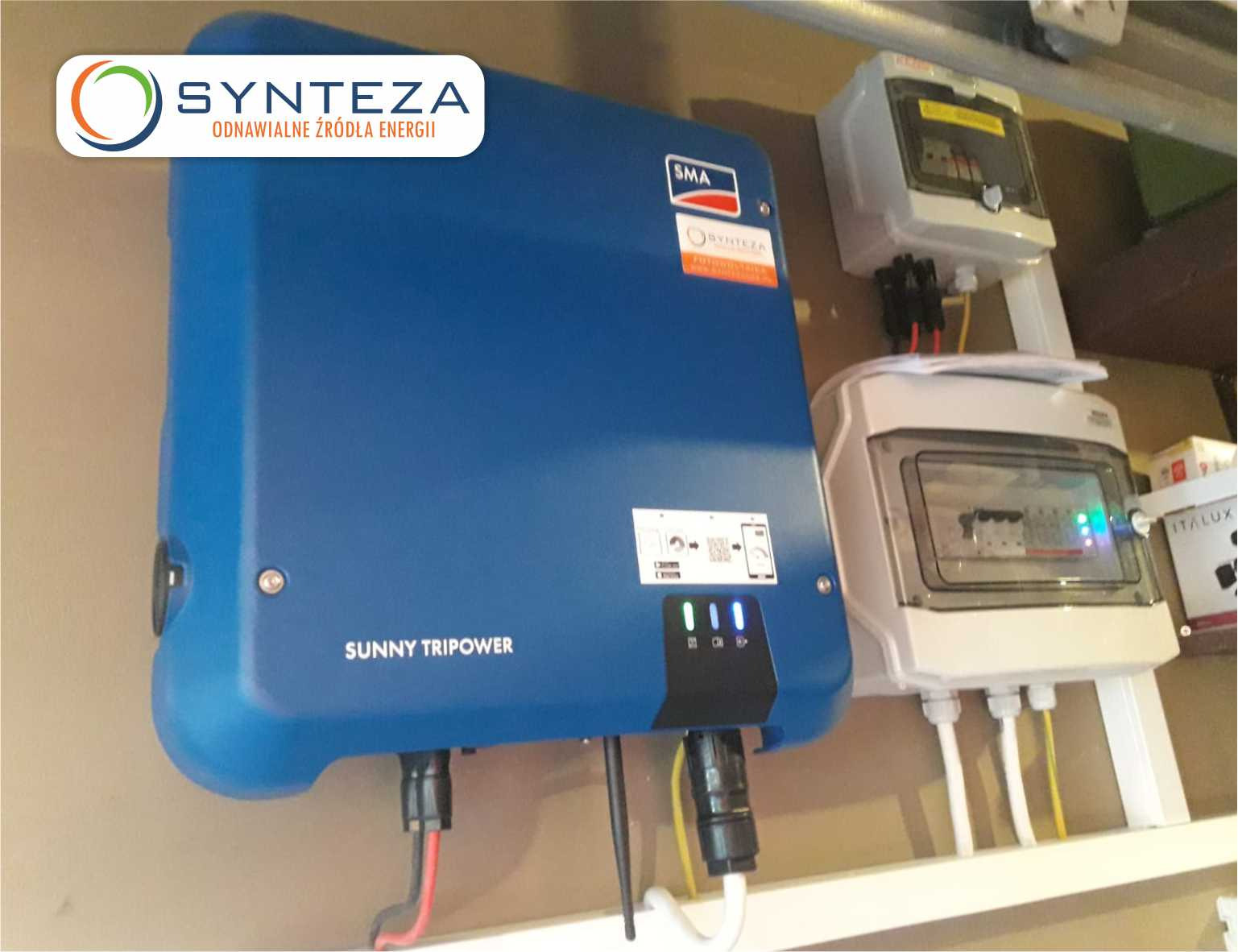 Niebieski falownik Sunny Tripower zamontowany na ścianie obok rozdzielnic elektrycznych z widocznymi przewodami.