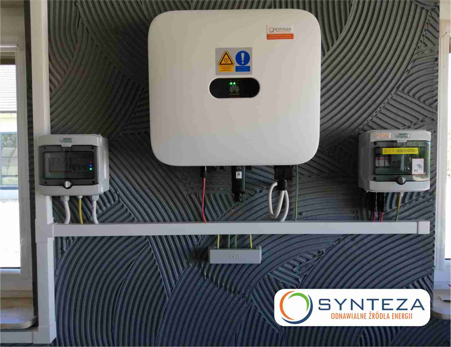 Biały inwerter fotowoltaiczny marki Huawei, zamontowany na ścianie o dekoracyjnej strukturze, z dwoma skrzynkami zabezpieczeń po bokach i logiem firmy Synteza w prawym dolnym rogu.