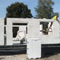 Mężczyzna w żółtej koszulce murowujący ścianę domu z białych bloczków, widoczne okna i fundament zabezpieczony czarną folią.