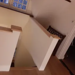 Widok z góry na odnowione schody z drewnianymi poręczami i świeżo pomalowanymi ścianami w jasnym odcieniu, z widocznym oknem na górze i fragmentem krzesła z ozdobnym oparciem w tle.