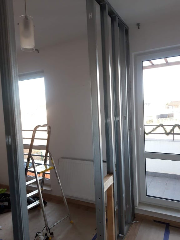 Wnętrze w trakcie budowy ścianki działowej z metalowych profili, widoczna drabina, grzejnik i okno balkonowe z dekoracją.