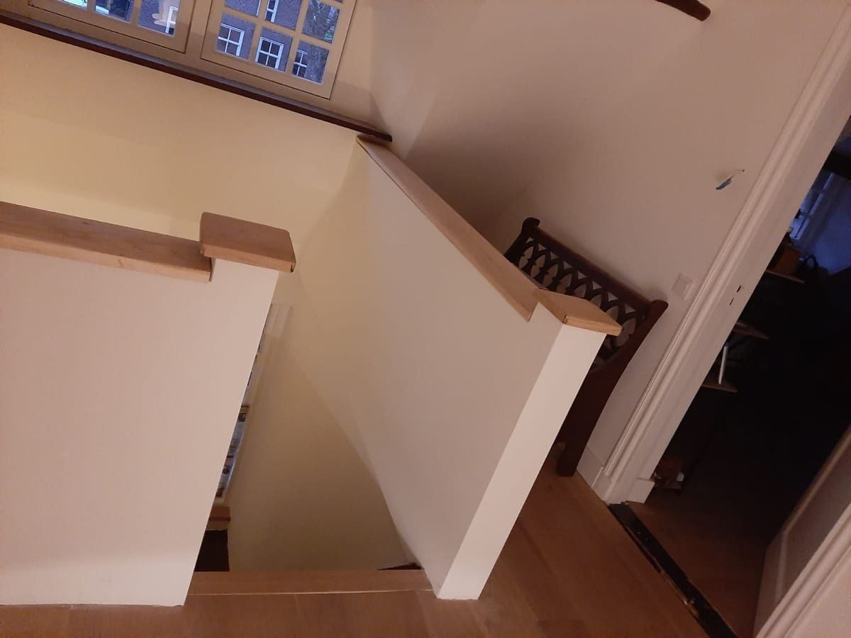 Widok z góry na odnowione schody z drewnianymi poręczami i świeżo pomalowanymi ścianami w jasnym odcieniu, z widocznym oknem na górze i fragmentem krzesła z ozdobnym oparciem w tle.