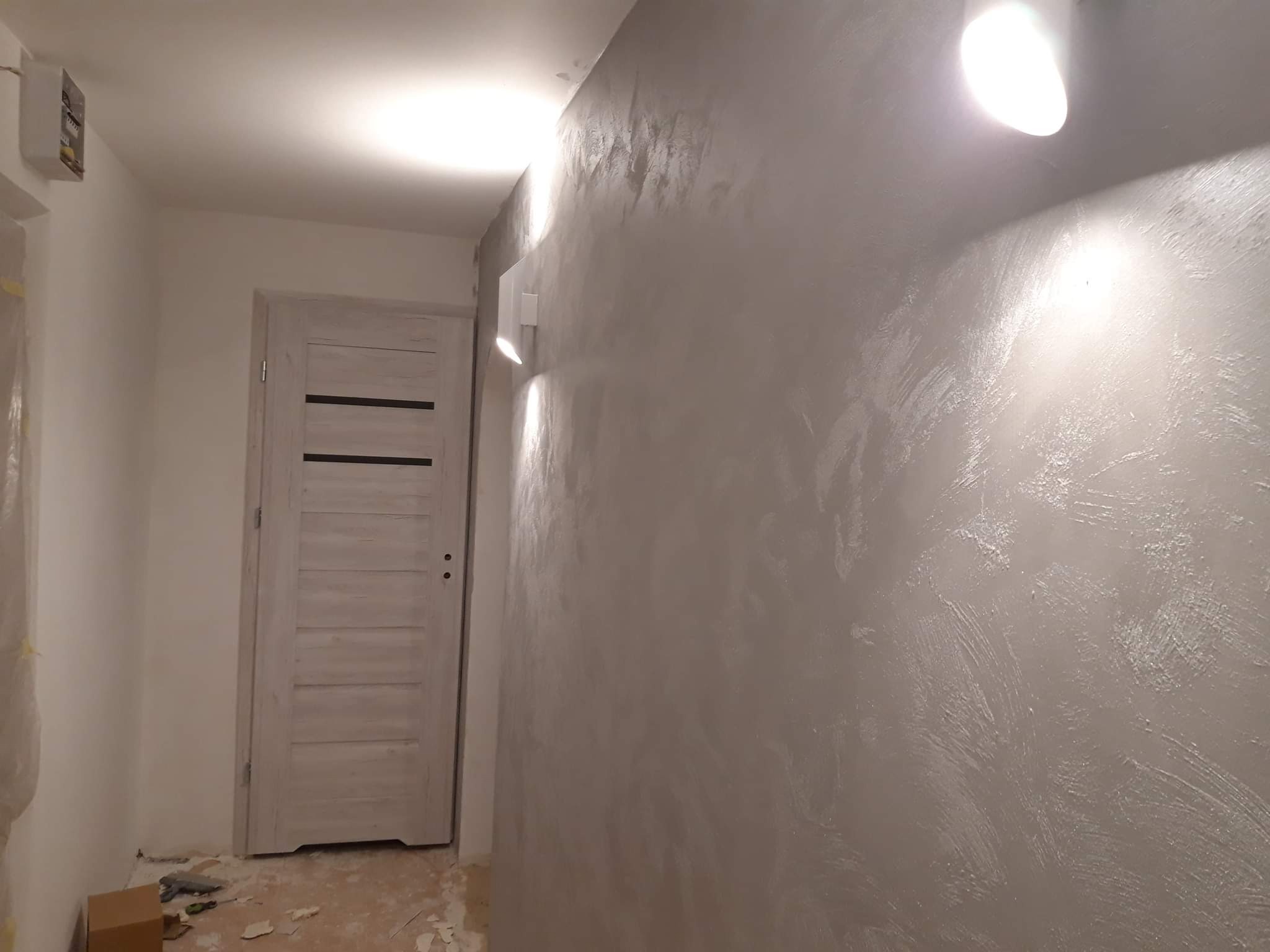 Ściana pokryta dekoracyjną szarą fakturą, oświetlona dwoma punktowymi lampami, widoczny fragment drzwi z poziomymi panelami i resztki po remoncie na podłodze.