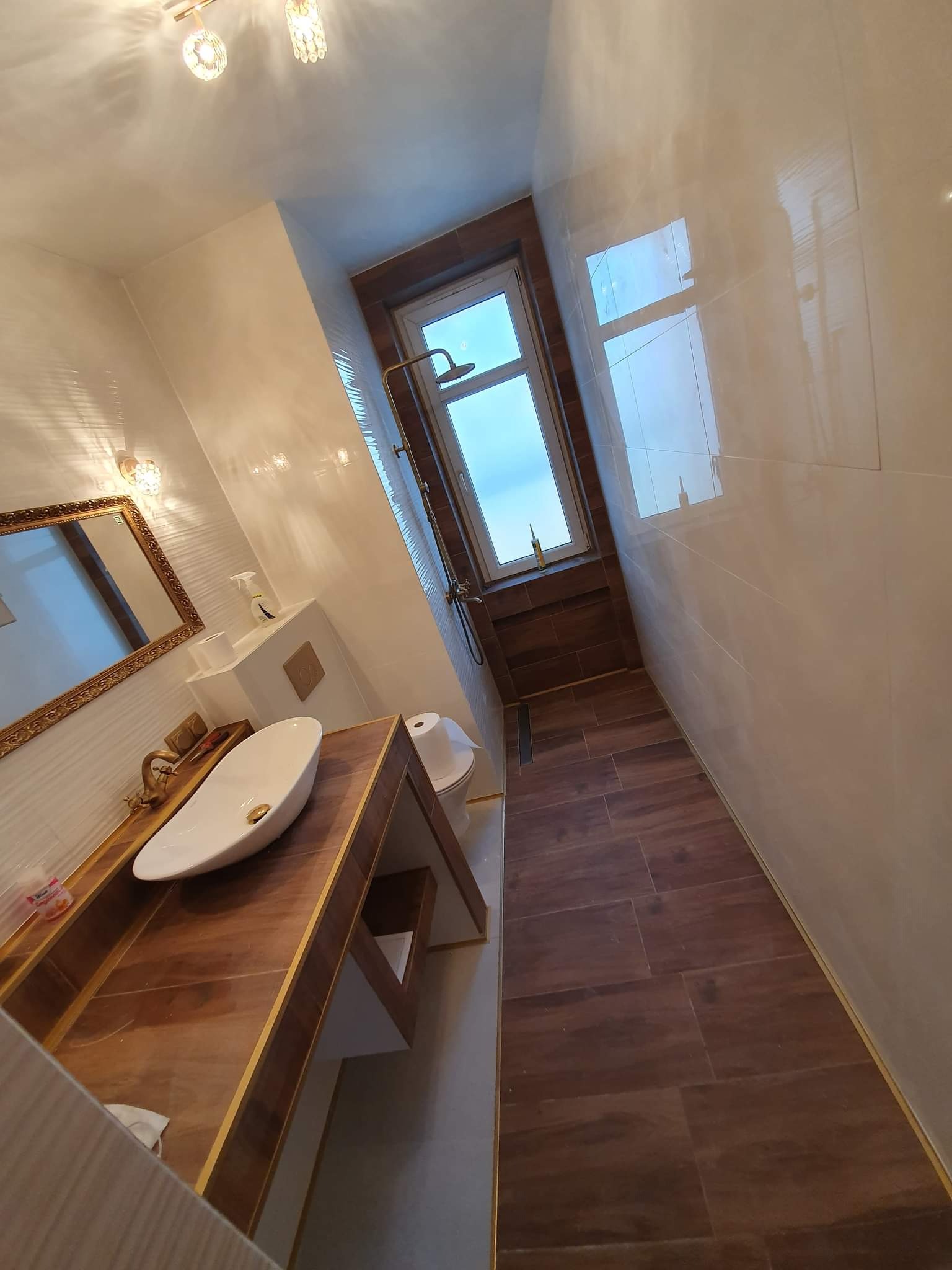 Nowoczesna łazienka z brązowymi płytkami podłogowymi imitującymi drewno, białą umywalką nablatową z złotymi akcentami, złotą armaturą i lustrem w bogato zdobionej złotej ramie, oświetlona przez...