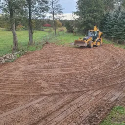 Żółta koparko-ładowarka na wyrównanym, brązowym terenie z wyraźnymi śladami opon, widoczne drzewa i ogrodzenie w tle, pochmurne niebo.