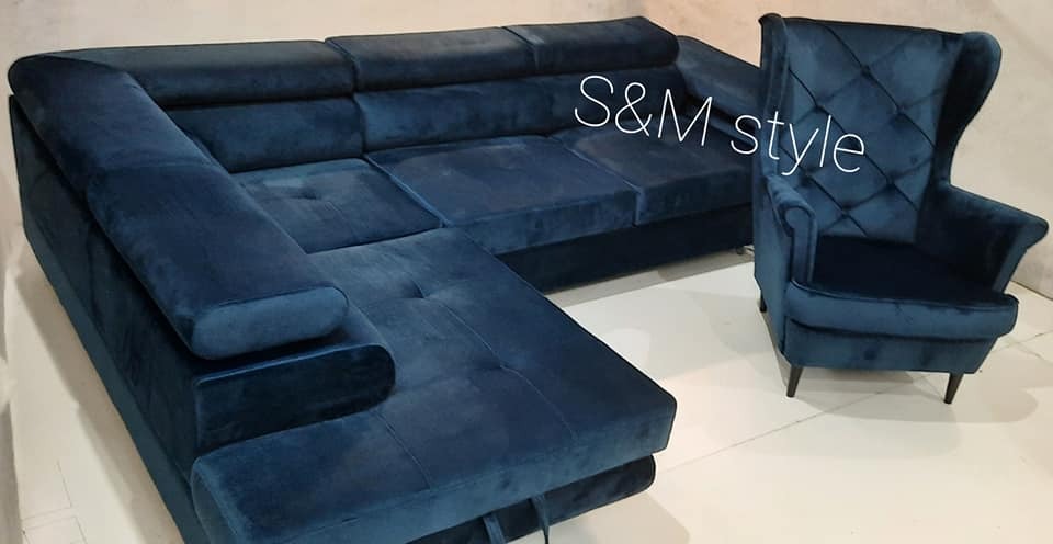 Granatowy narożnik tapicerowany welurem z funkcją spania i fotel uszak w tym samym stylu, z widocznym logo firmy na narożniku.
