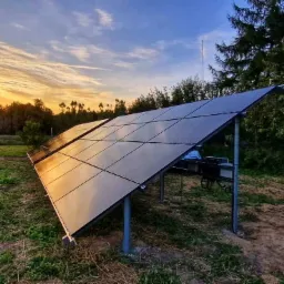 Instalacja paneli słonecznych na metalowej konstrukcji, odbijających zachodzące słońce na tle trawnika i drzew