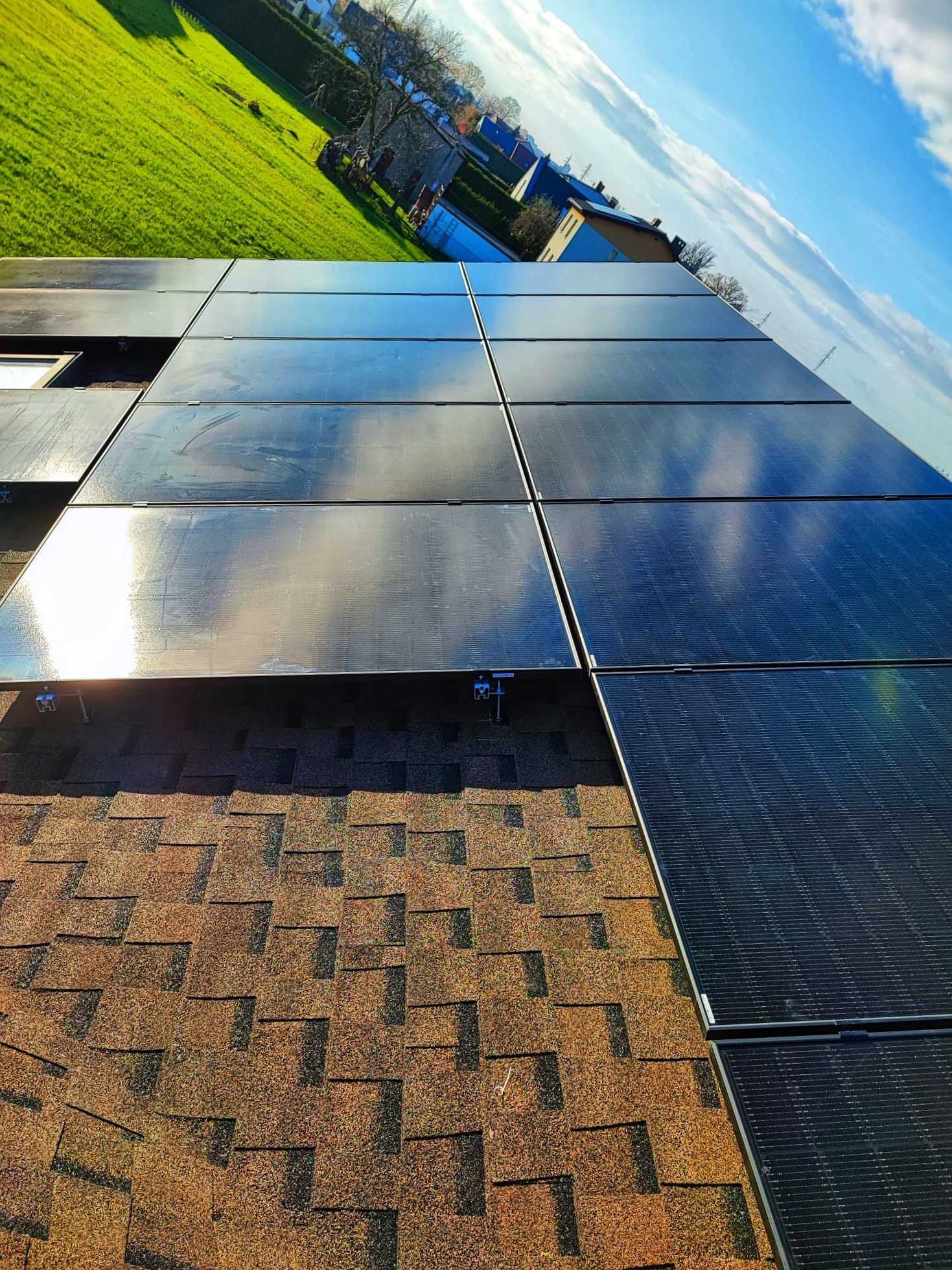 Czarny panel słoneczny zamontowany na brązowym dachu z gontów bitumicznych, widok na zielony trawnik i domy w tle, błękitne niebo z chmurami odbija się w panelach.