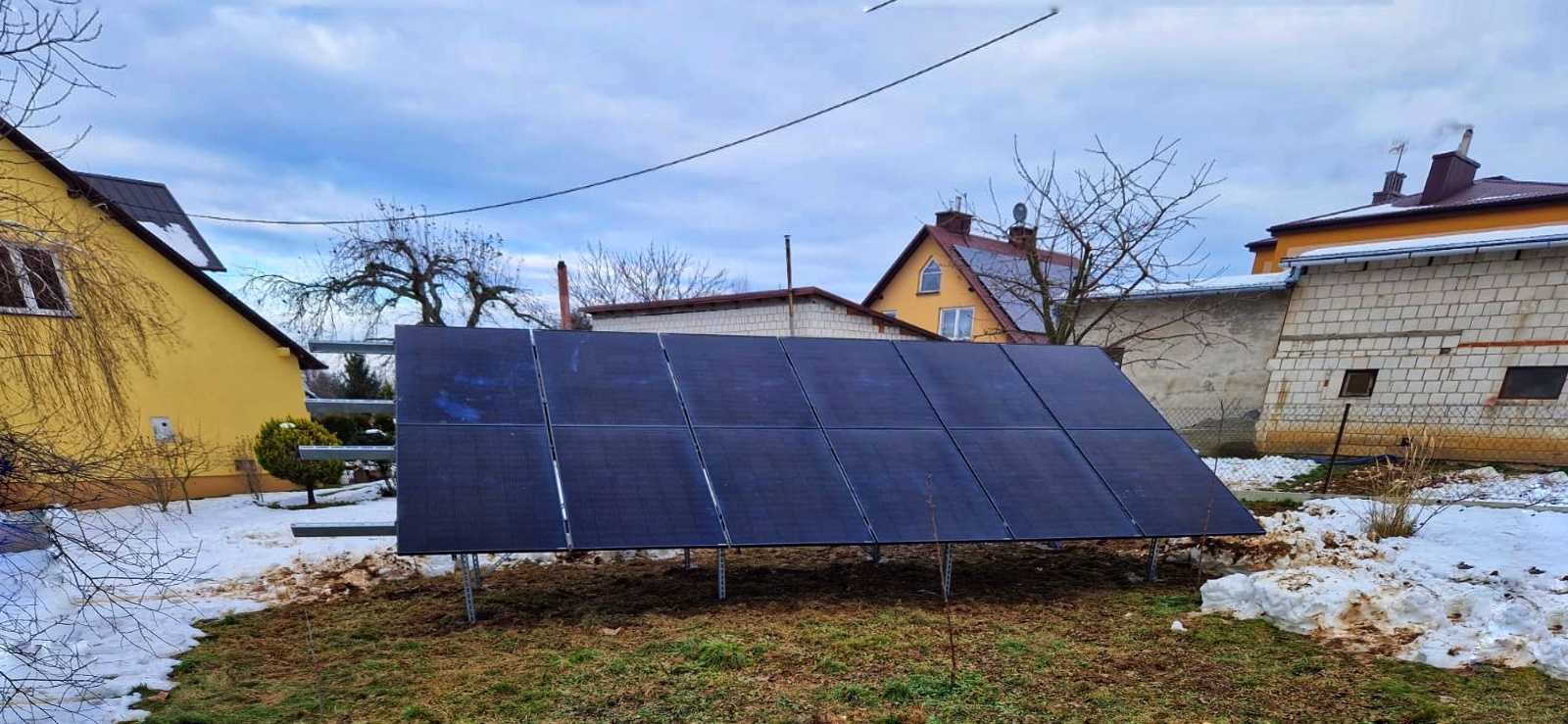 Panel słoneczny ustawiony na metalowej konstrukcji na trawniku z resztkami śniegu, na tle domy z dachami pokrytymi śniegiem i błękitne niebo.