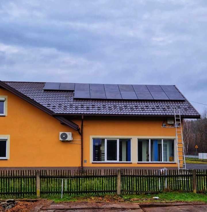 Żółty dom jednorodzinny z ciemnymi panelami fotowoltaicznymi na dachu z dachówki, drabina oparta o ścianę, widoczny klimatyzator na elewacji.