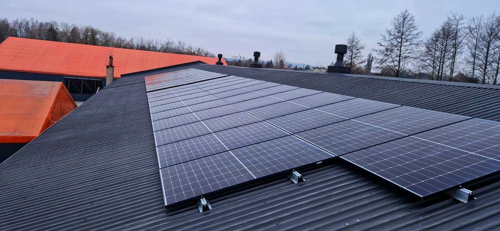 Instalacja paneli słonecznych na falistym, metalowym dachu budynku w Niechobrzu, widok z góry, z kominami w tle i fragmentem pomarańczowego dachu innego budynku.