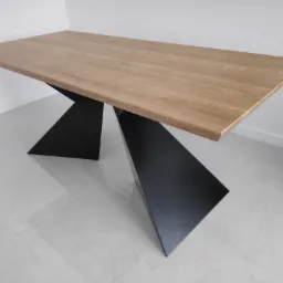 Stół z blatem z jasnego drewna i geometryczną, czarną podstawą na tle białej ściany i podłogi z jasnych płytek.