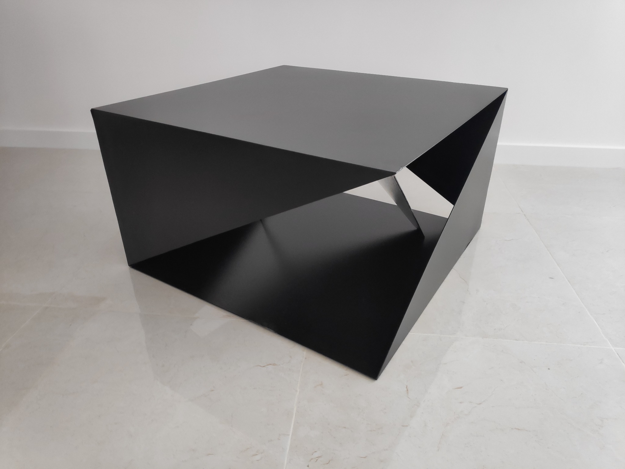 Czarny, geometryczny stolik kawowy o konstrukcji przypominającej sześcian z wyciętymi trójkątnymi fragmentami, ustawiony na jasnej, marmurowej podłodze przy białej ścianie.