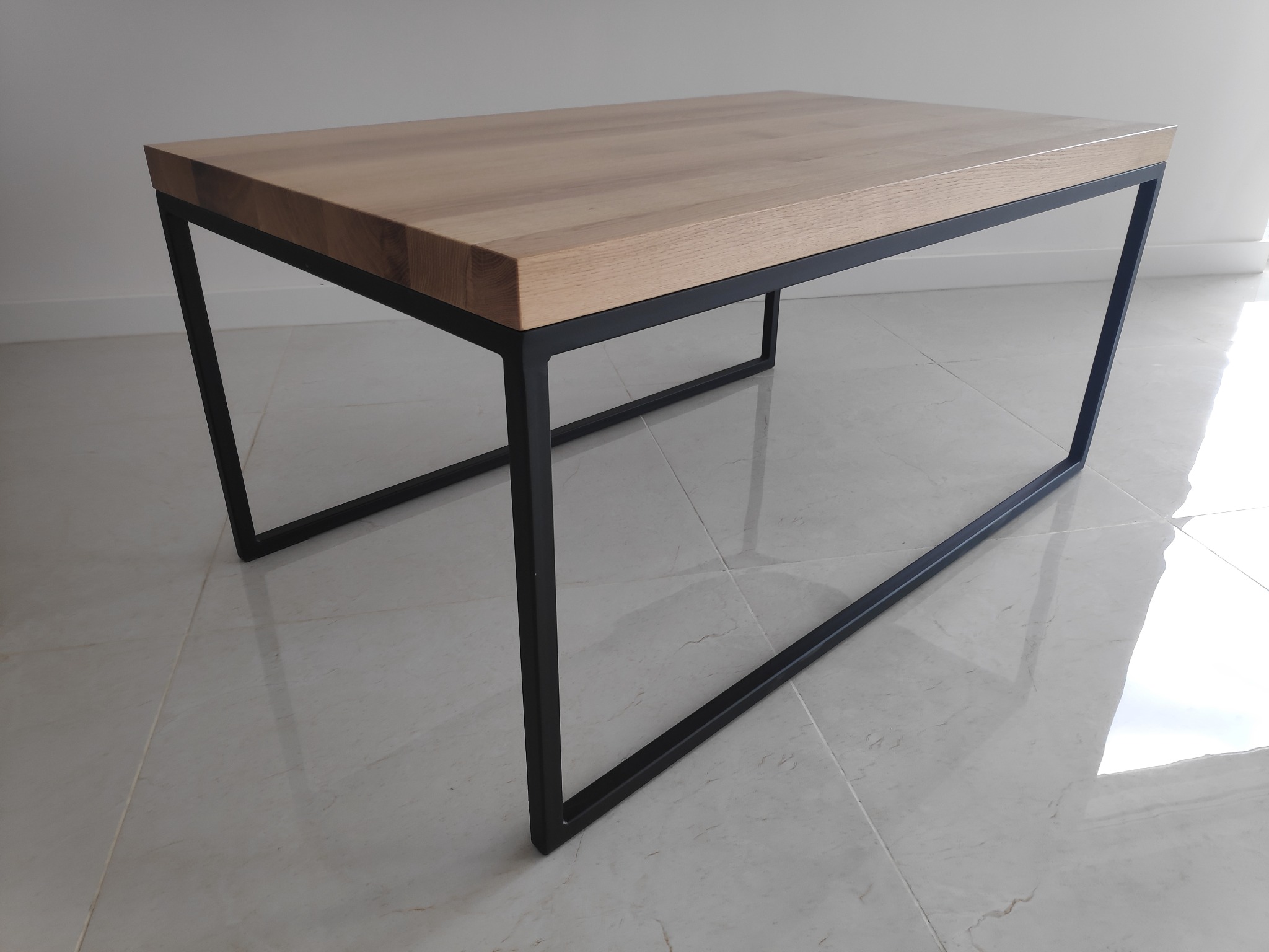 Minimalistyczny stolik kawowy z jasnego drewna na czarnych, metalowych nogach, wykonany w Rumi, na tle białej ściany i błyszczącej, jasnej podłogi.