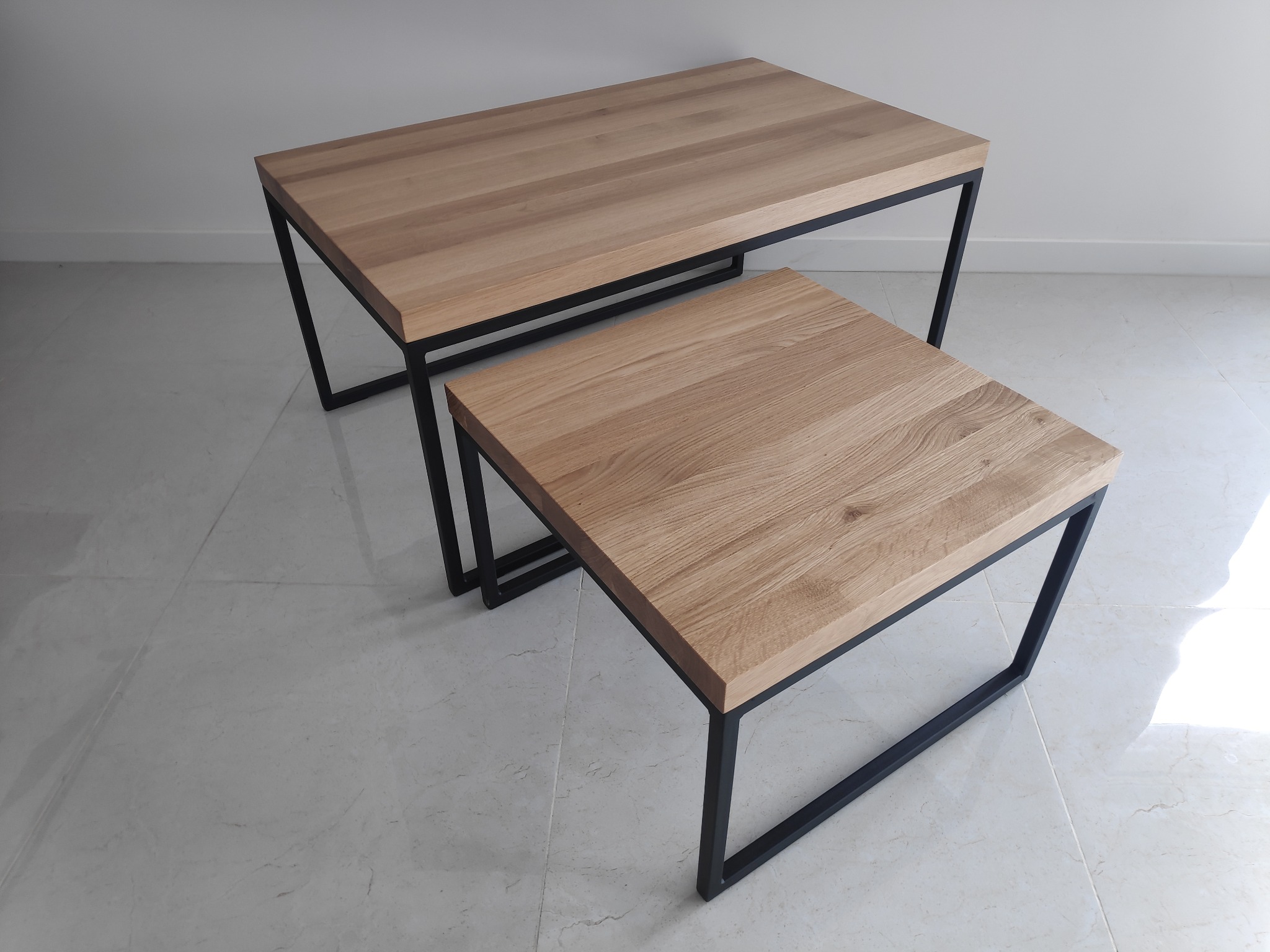 Dwa stoliki kawowe z drewnianymi blatami i czarnymi, metalowymi nogami, ustawione na marmurowej podłodze na tle białej ściany.