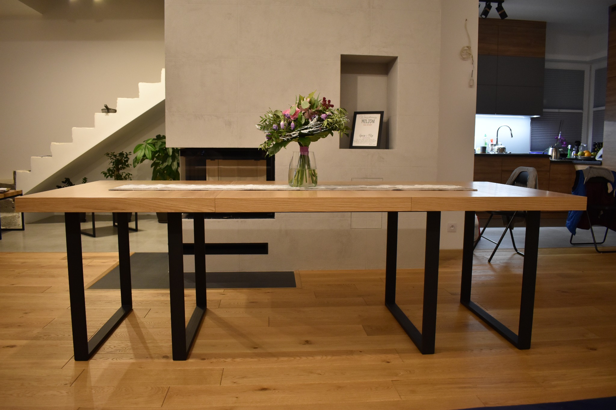 Jasny, drewniany stół z czarnymi nogami w nowoczesnym wnętrzu z drewnianą podłogą, na stole wazon z bukietem kwiatów, w tle schody i kominek.