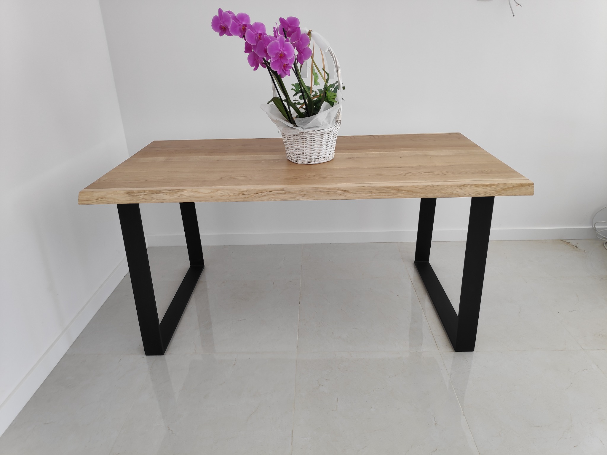 Prostokątny stół z jasnego drewna z wyraźnym usłojeniem, osadzony na czarnych, metalowych nogach w kształcie litery V, na blacie biały koszyk z fioletowymi storczykami, całość na tle białej ściany...