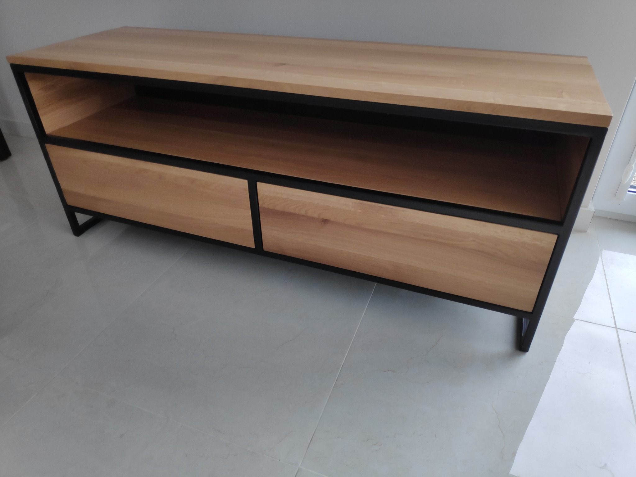 Stolik RTV z jasnego drewna z czarną metalową ramą, dwie szuflady i otwarta półka, na białej podłodze z płytek.