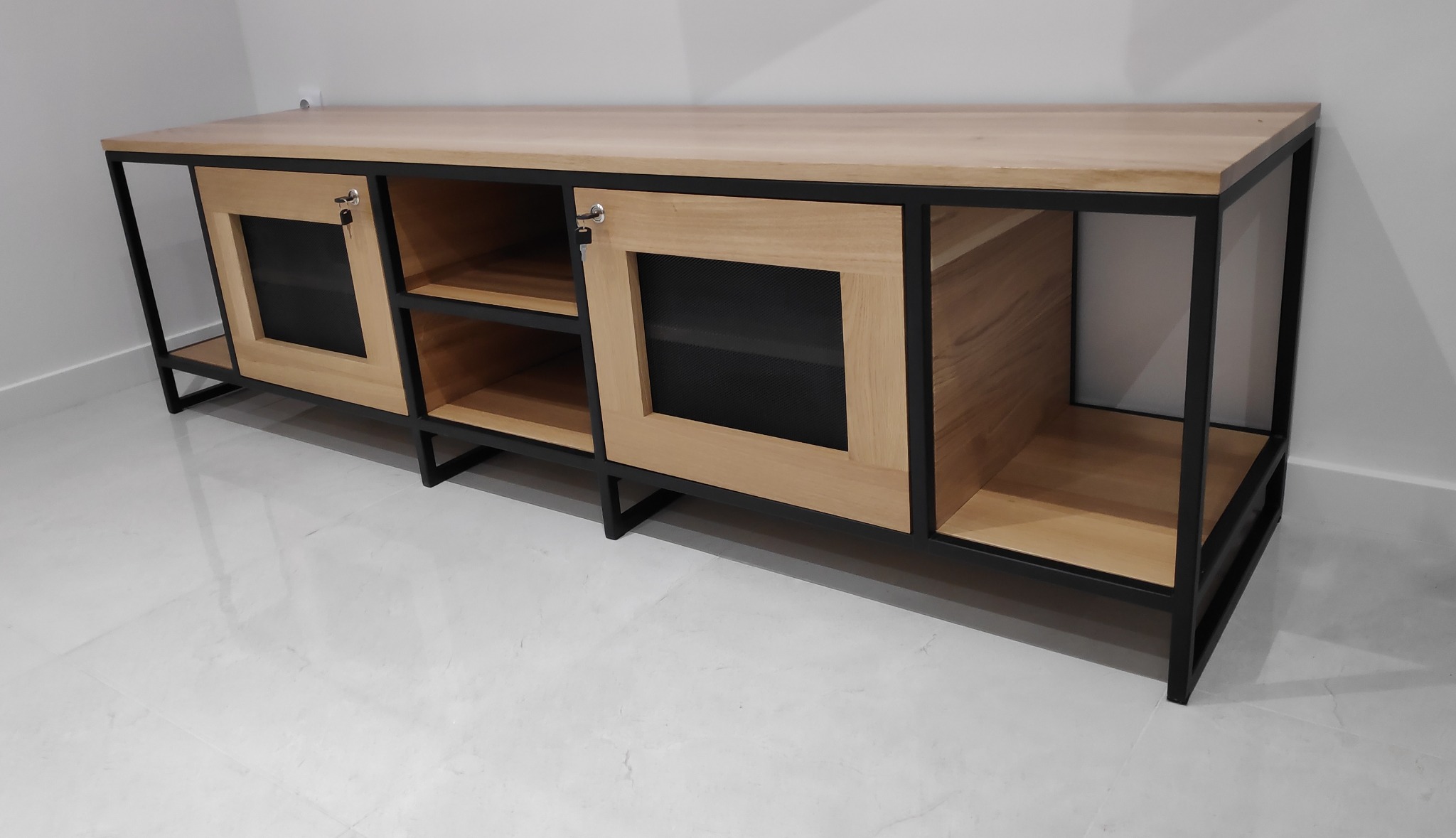 Długa komoda z jasnego drewna z czarną metalową ramą, zamykana na klucz, na tle białej ściany i podłogi.