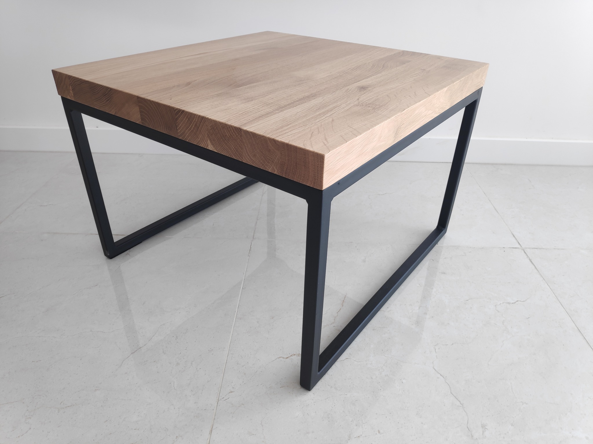 Kwadratowy stolik kawowy z blatem z jasnego drewna i czarną, minimalistyczną podstawą z metalowych profili, ustawiony na jasnej marmurowej podłodze.