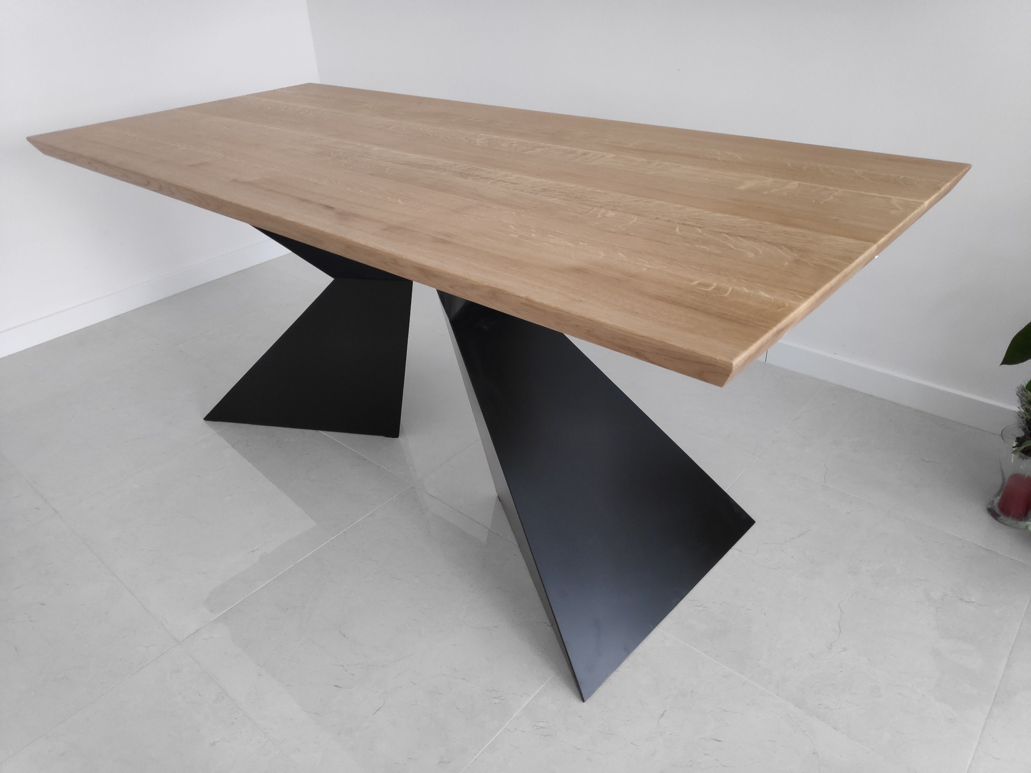 Stół z blatem z jasnego drewna i geometryczną, czarną podstawą na tle białej ściany i podłogi z jasnych płytek.