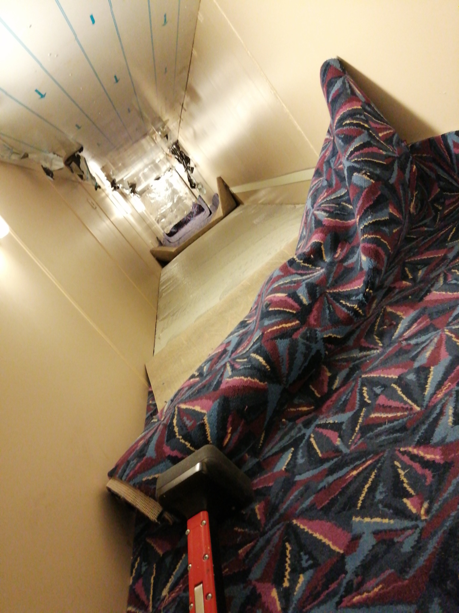 Fragment wnętrza z częściowo zamontowaną wykładziną o wzorze geometrycznym i narzędziem do jej naciągania, widoczne ocieplenie sufitu i ściany.