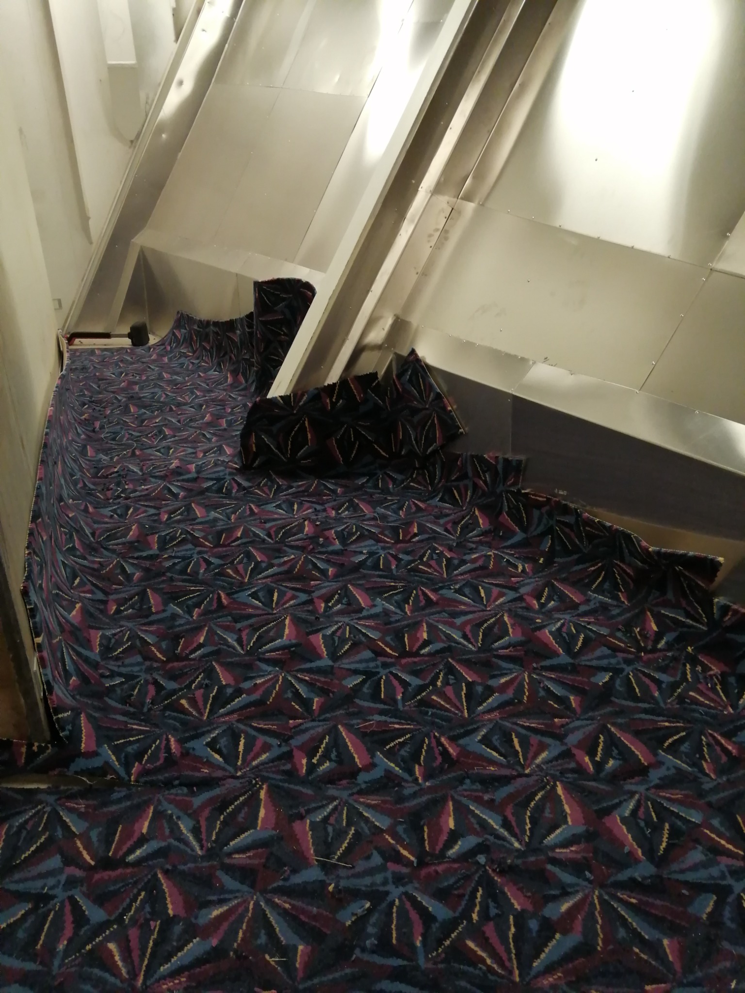 Fragment montażu wzorzystej wykładziny dywanowej w nietypowej przestrzeni z metalowymi ścianami, widoczne niedociągnięte krawędzie i przycięte kawałki.