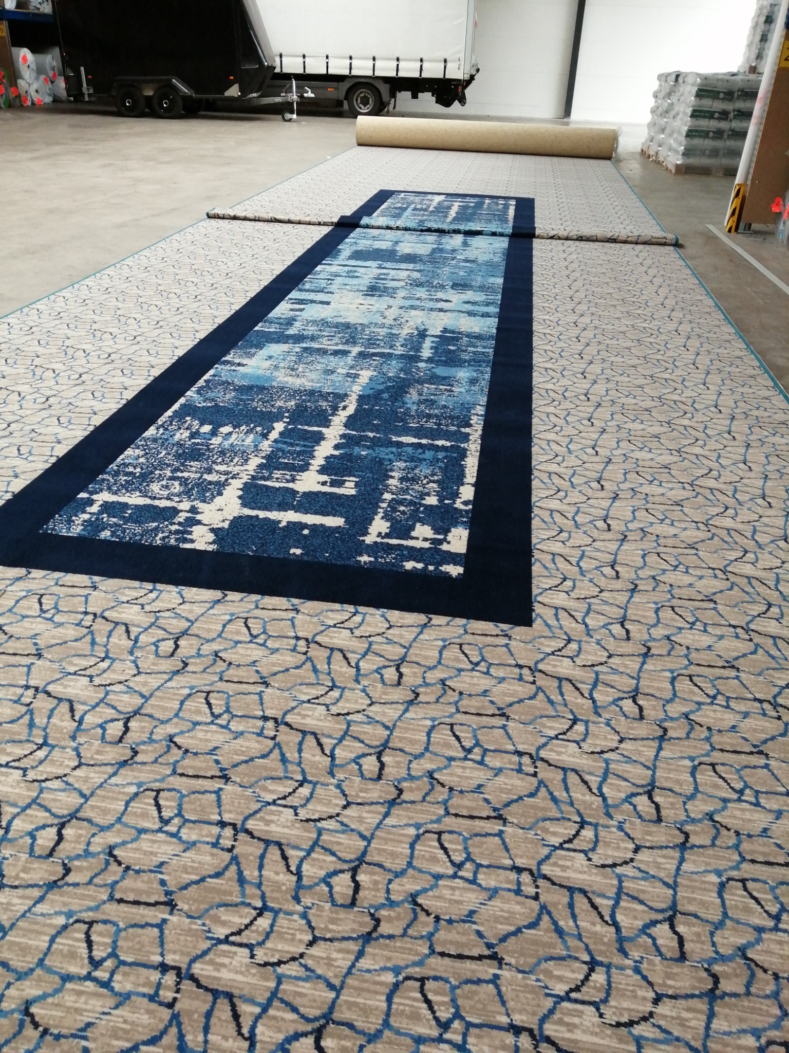 Wielkoformatowa wykładzina dywanowa z geometrycznym wzorem i niebieskim pasem pośrodku, częściowo rozwinięta, z widocznym transportem w tle.