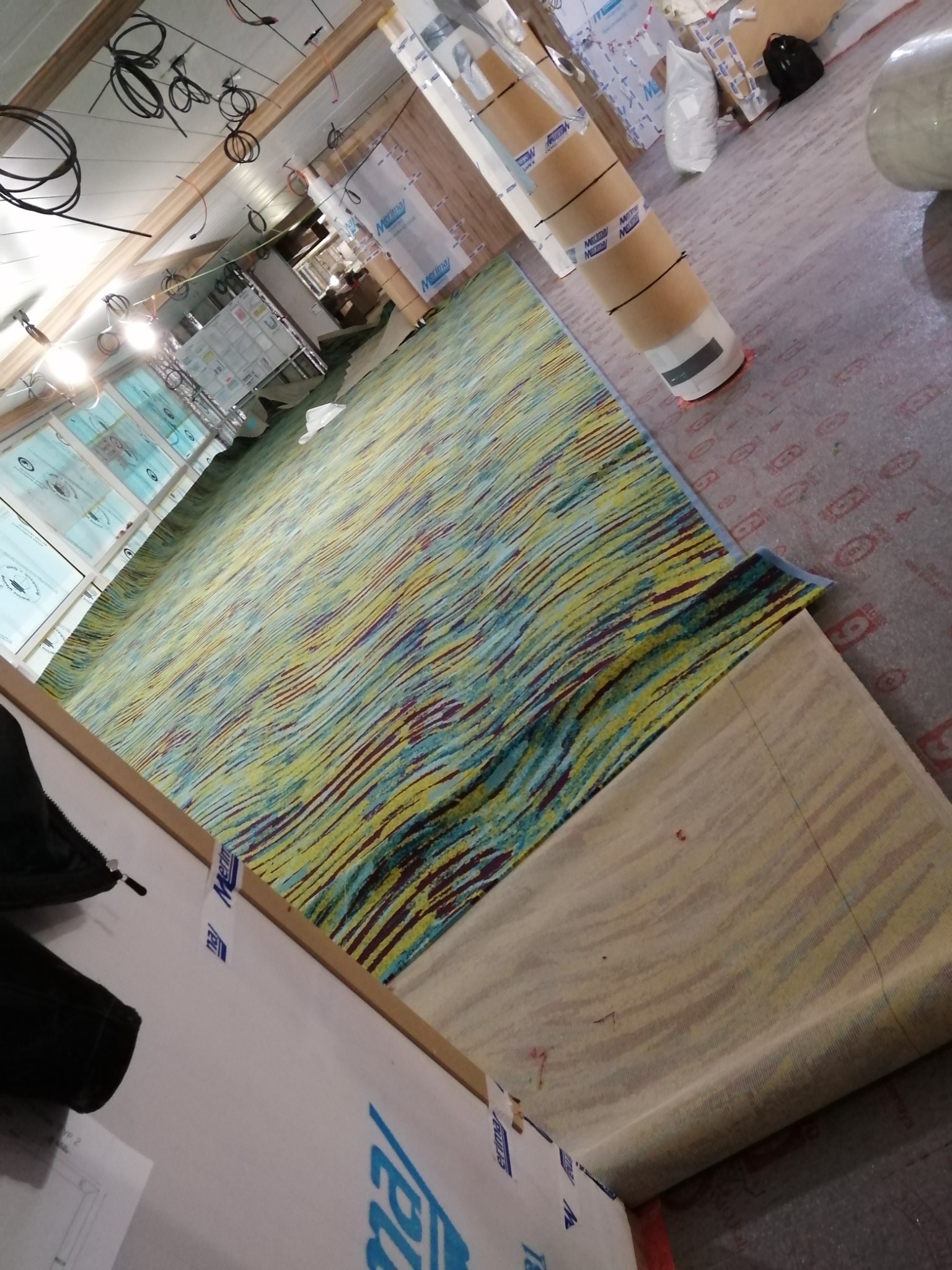 Rozwinięty fragment wzorzystej wykładziny dywanowej na podkładzie, widoczne rolki materiału i instalacje pod sufitem w trakcie prac wykończeniowych.