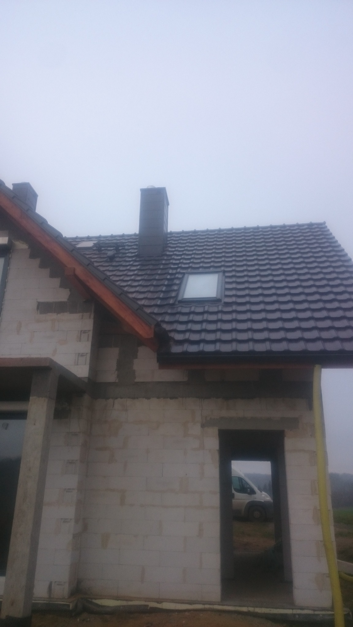 Surowy stan domu w trakcie budowy, widoczna ściana z pustaków, dach z ciemnej dachówki z oknem dachowym, komin i częściowo widoczny samochód przez otwór drzwiowy.