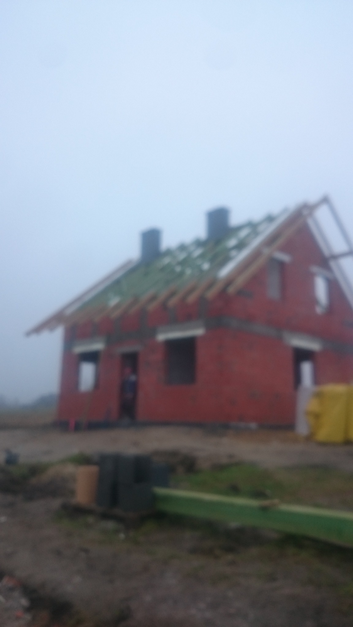 Surowy stan domu z czerwonej cegły w mglisty dzień, widoczne kominy, konstrukcja dachu z zielonymi elementami, teren budowy w nieostrym planie.