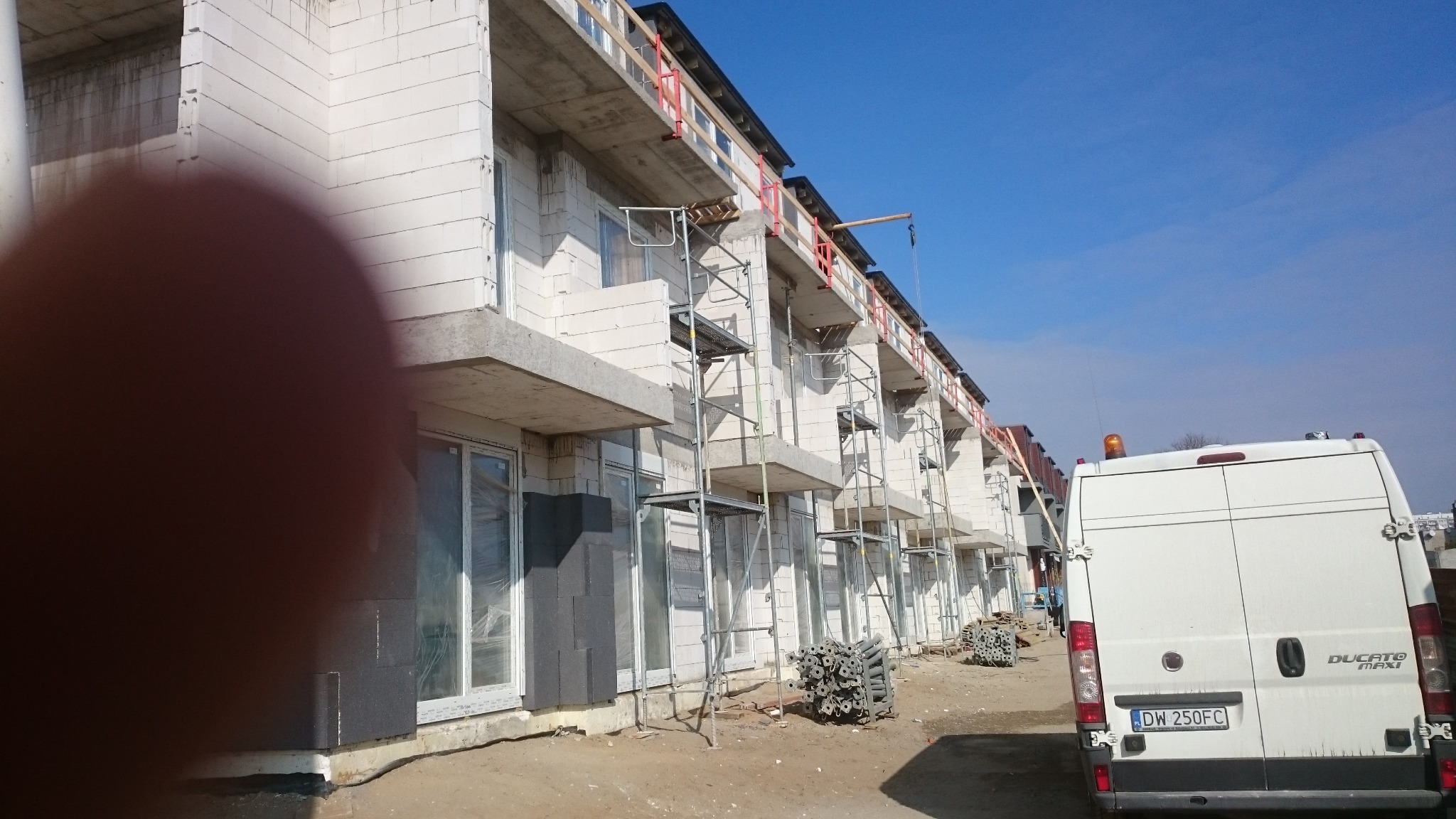 Wznoszenie szeregu budynków z białego betonu komórkowego, widoczne rusztowania, zabezpieczenia na balkonach, częściowo ocieplone ściany, biały samochód dostawczy i palety ze stalowymi elementami...