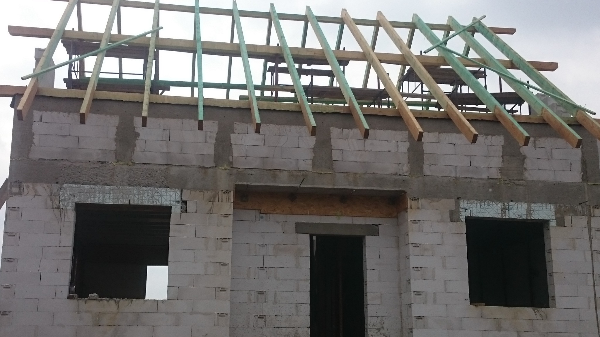 Surowy stan domu murowanego z bloczków betonowych, widoczne ściany, otwory okienne z zabezpieczeniem, drewniana konstrukcja dachu w trakcie budowy, rusztowanie w tle.