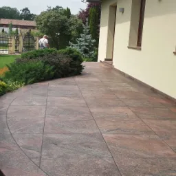 Wyłożony brązowo-szarymi płytkami taras kamienny przylegający do jasnej elewacji budynku z dwoma oknami, z widocznym fragmentem ogrodu i postacią w tle.