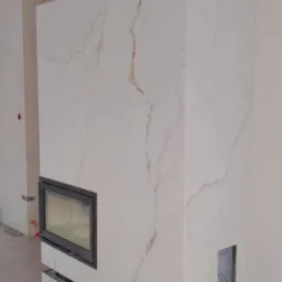 Obudowa kominka z białego marmuru z ciemnymi szczelinami wentylacyjnymi i wkładem kominkowym; podłoga w trakcie wykańczania.