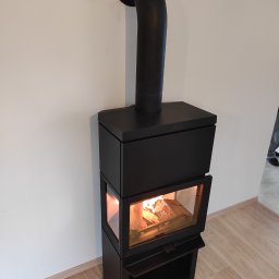 P.P.H.U. Stone-Service Artur Tomkowiak - Czarny, nowoczesny piec wolnostojący z widocznym płomieniem, podłączony do komina, na tle jasnej ściany i podłogi imitującej drewno.