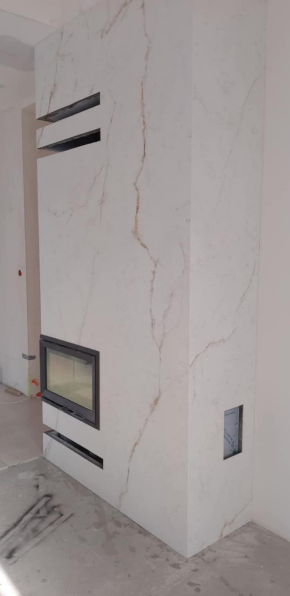 Obudowa kominka z białego marmuru z ciemnymi szczelinami wentylacyjnymi i wkładem kominkowym; podłoga w trakcie wykańczania.