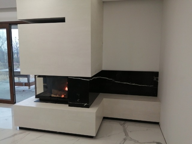 Nowoczesny kominek z białym obramowaniem i czarnym kamiennym akcentem, widoczne palenisko z płomieniami, marmurowa podłoga, minimalistyczny design.