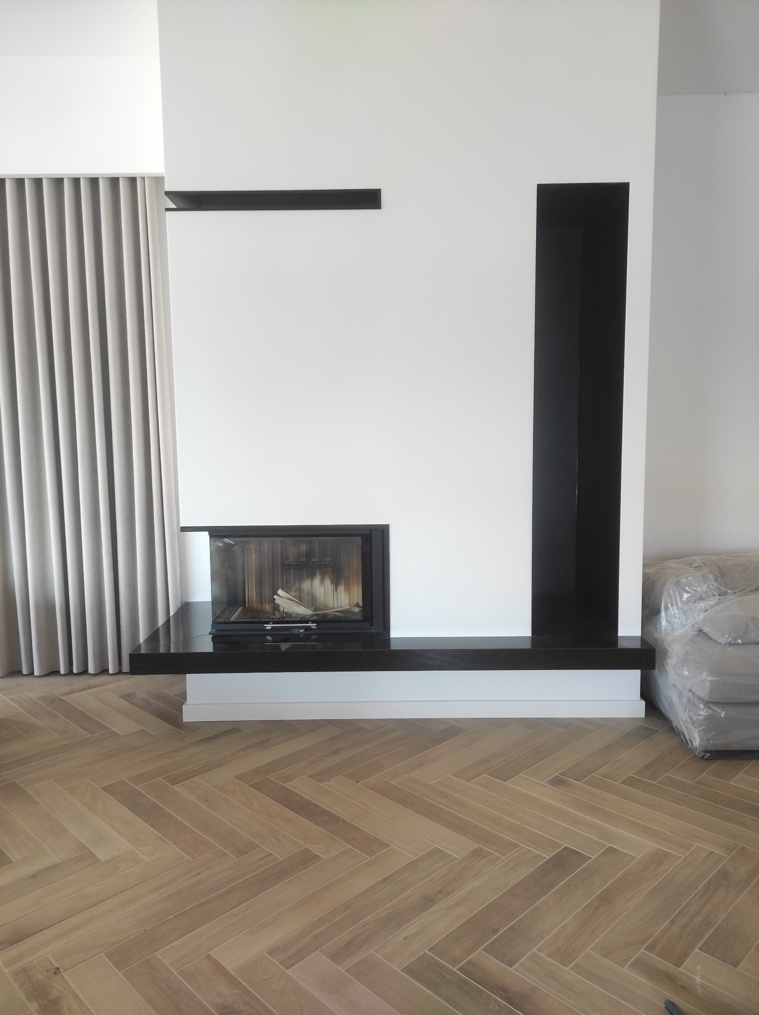 Nowoczesny kominek z czarnym obramowaniem i blatem z ciemnego kamienia, wbudowany w białą ścianę z minimalistycznymi półkami, na podłodze parkiet w jodełkę.