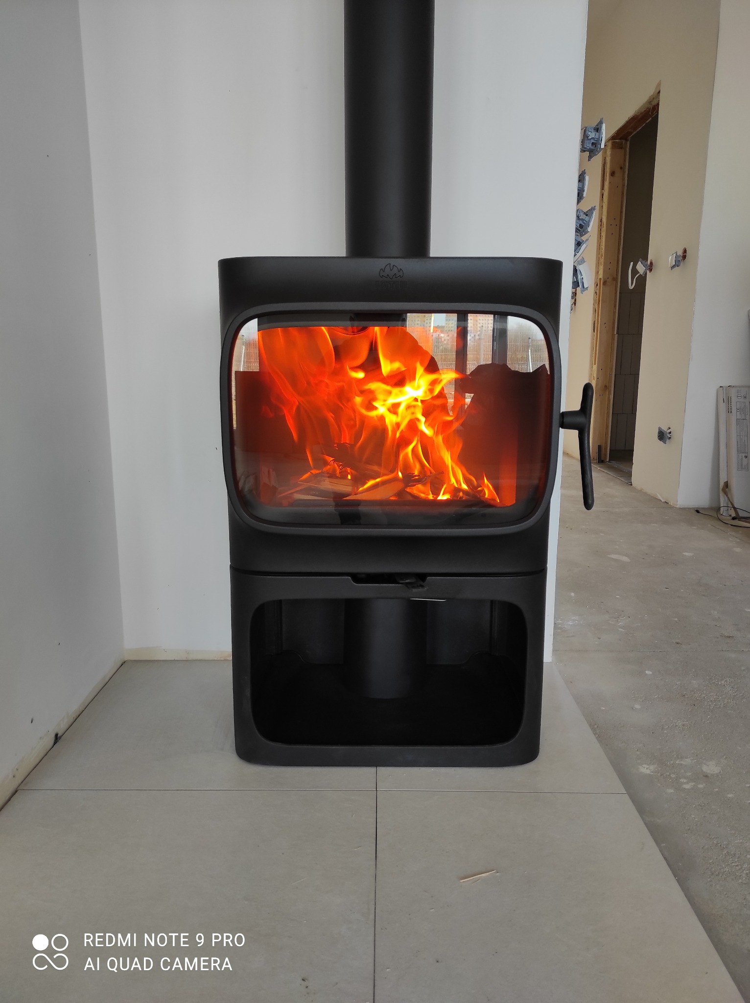 Nowoczesny, czarny piec wolnostojący z płonącym ogniem w szklanej obudowie, umieszczony na jasnej, kwadratowej posadzce w surowym wnętrzu z widocznymi elementami instalacji elektrycznej.