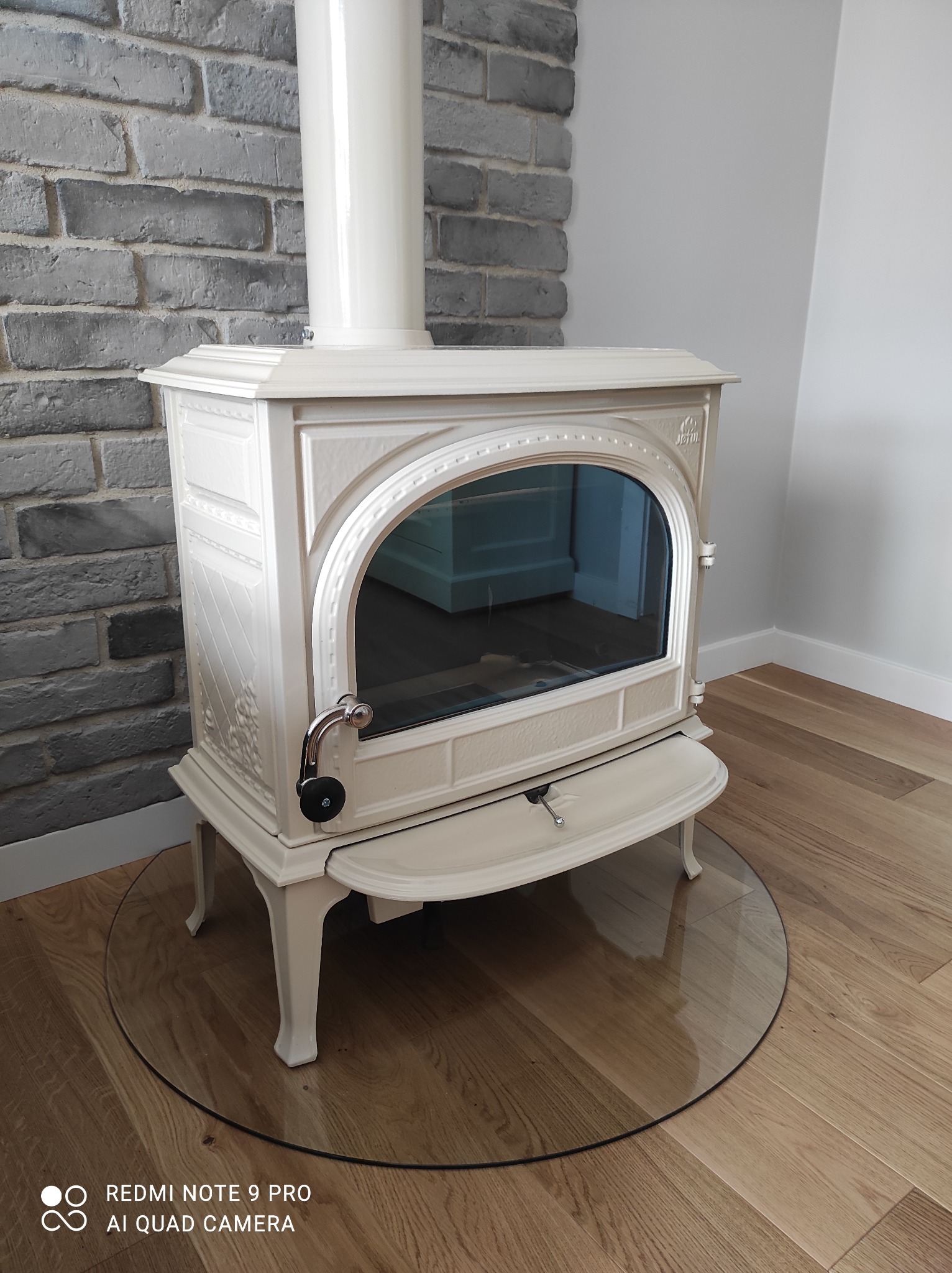 Elegancki, kremowy piec wolnostojący z szybą, ustawiony na szklanej podstawie na drewnianej podłodze, z szarą cegłą dekoracyjną w tle.