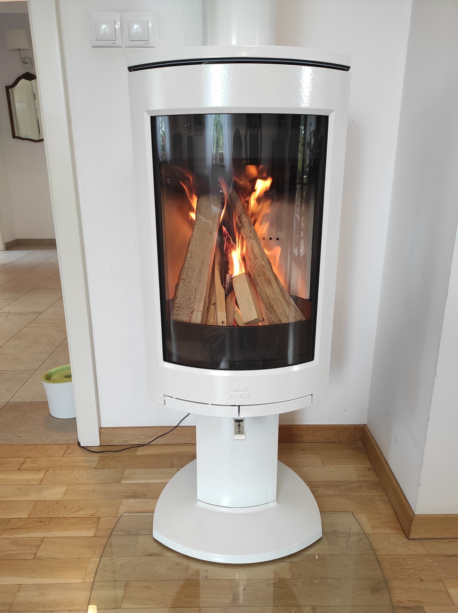 Nowoczesny, wolnostojący, biały piec kominkowy z palącym się drewnem w szklanej obudowie, na tle jasnego wnętrza z parkietem.