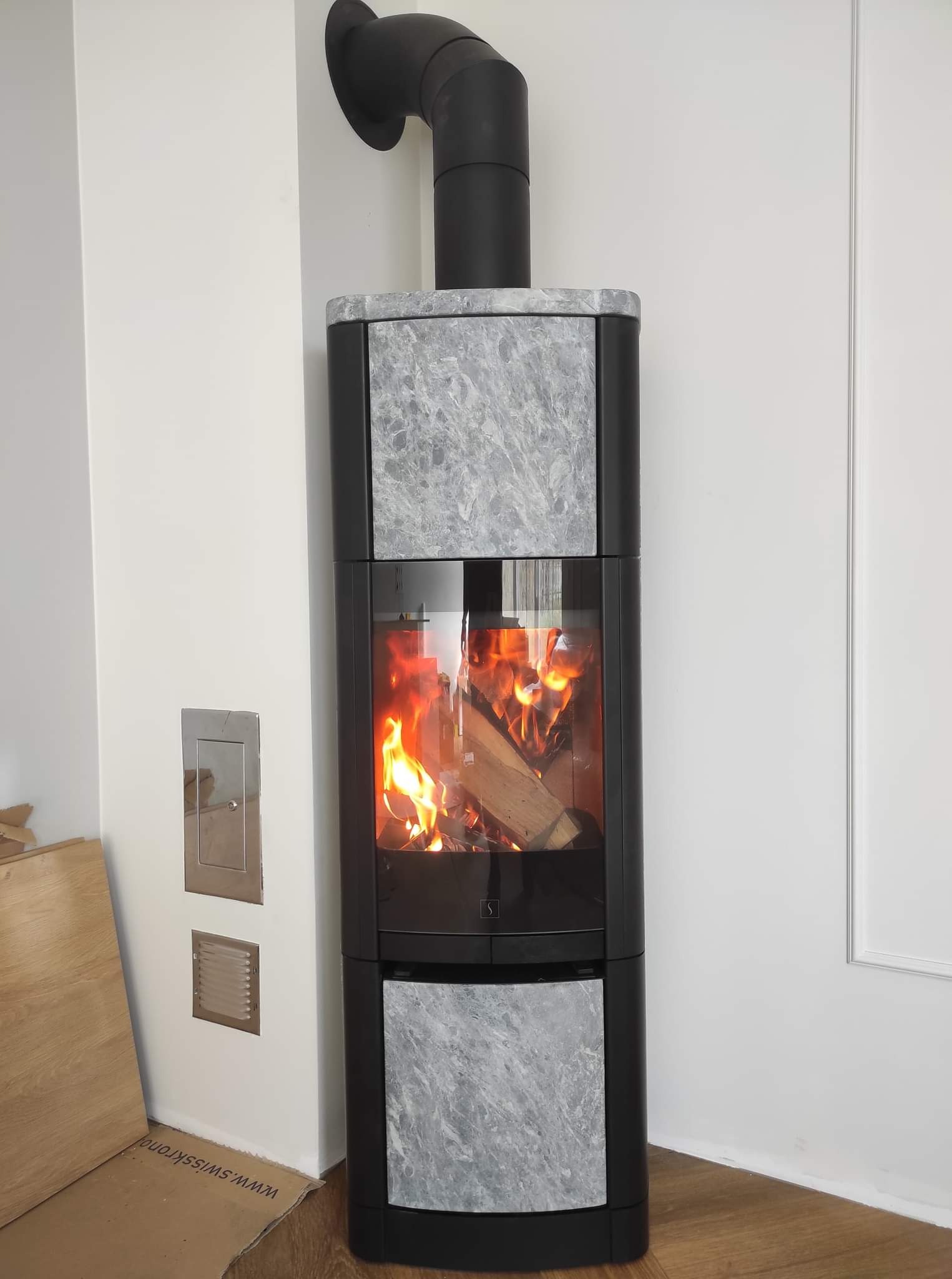 Nowoczesny piec wolnostojący z elementami wykończeniowymi z szarego kamienia, palenisko z płonącym drewnem, czarny komin odprowadzający dym, widoczne elementy instalacji na ścianie.