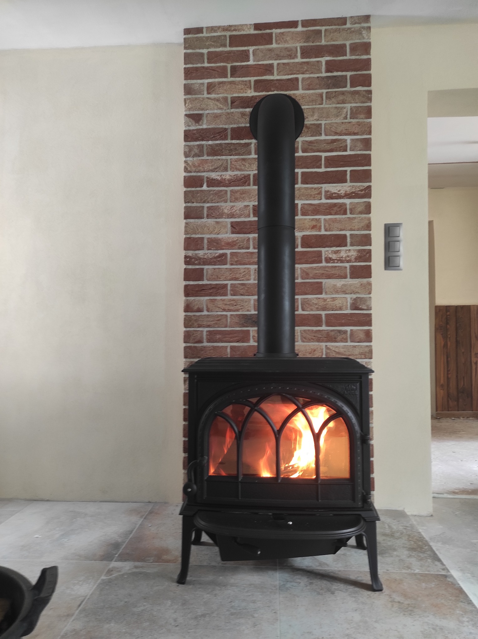 Czarny, żeliwny kominek z płomieniami wewnątrz, umieszczony na tle ściany z cegły i jasnej ściany, z widoczną podłogą z jasnych płytek i fragmentem pomieszczenia w tle.