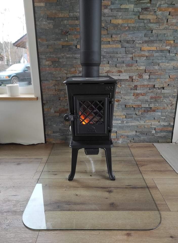 Żeliwny piec wolnostojący marki Jotul model 602 z płomieniami widocznymi przez ozdobne drzwiczki, ustawiony na szklanej płycie ochronnej na drewnianej podłodze, z tłem z kamienia dekoracyjnego.