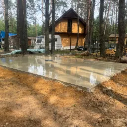 Świeżo wylany fundament pod budynek w lesie, widoczne odbicia drzew w mokrym betonie.